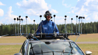 A photo of someone in a blue jacket with their upper body through the sunroof of a car. There is a ring of several small microphones surrounding them.