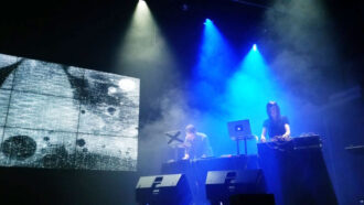 a woman and a man on a soundstage, both are using laptops and audio mixers to create a dance music performance. The room is dark and smoky, performers are lit by beams of blue light.