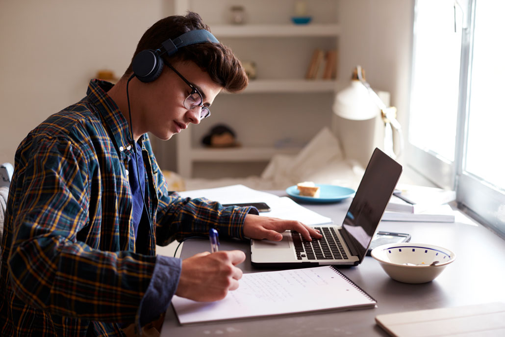 a view of a young man wearing headphones, sitting at a desk, and focusing on his school work. He's taking notes and using a laptop.