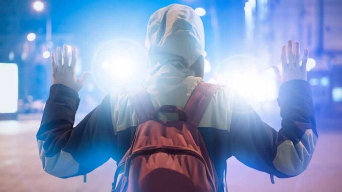 a person facing wearing a hoody and a backpack is standing with their hands in the air facing away from the camera and toward bright lights