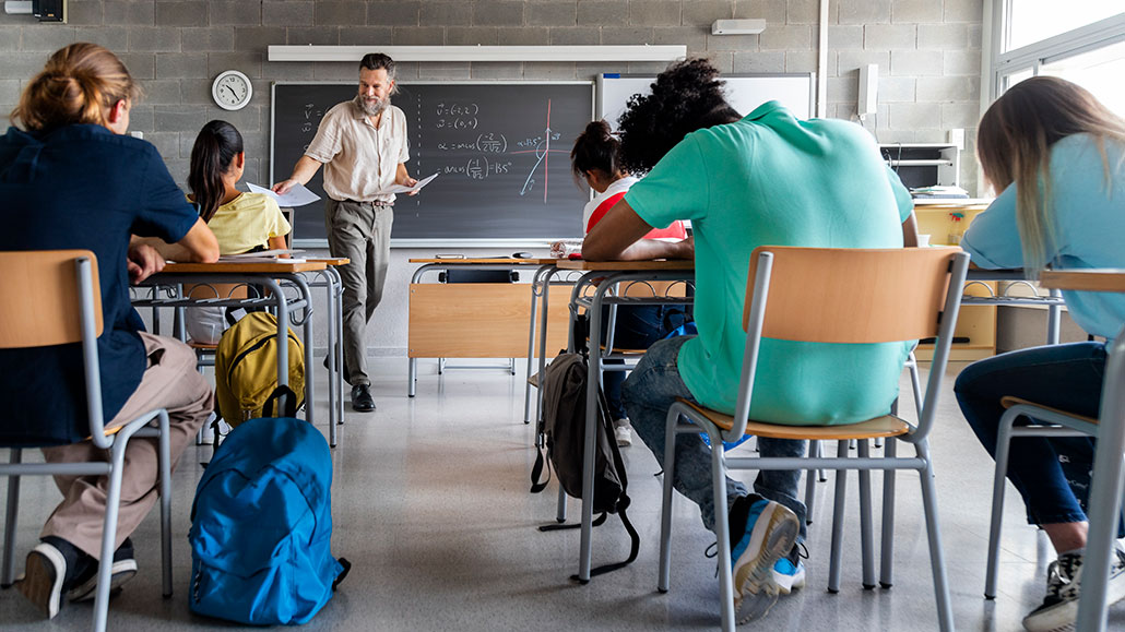 a photo of a classroom with student facing forward. A teacher is at the front handing out assignments