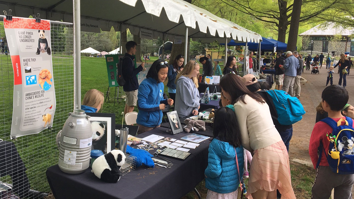 a woman stands at a booth on the sidewalk of a park and talks to parents and their kids