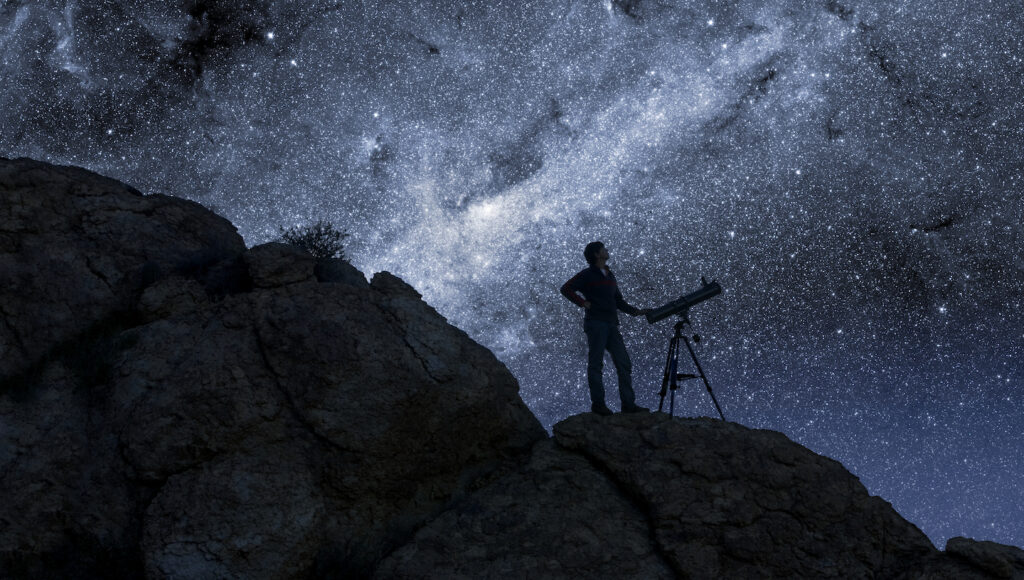 A person with a telescope standing on a dark hillside, silhouetted against a starry night sky.