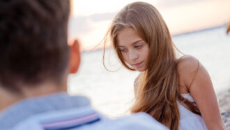 a sad young woman with long blond hair can be seen over the shoulder of person facing away from the viewer