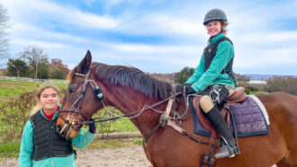 a blond 12-year old girl wearing equestrian gear sits on a horse while her twin sister stands near the horse's head, holding its bridle