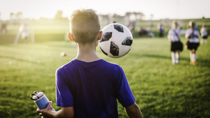 a sweaty kid on a soccer field is holding a water bottle. A soccer ball is floating in front of him.