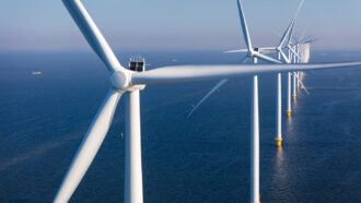 An aerial photo of a line of offshore wind turbines rising above a dark blue ocean. Ships can be seen in the distance.