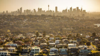 an aerial photo of Sydney, Australia on a hot and hazy day. At the bottom of the image are houses perched on a sea cliff. Towards the top of the picture the Sydney skyline can be seen.