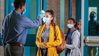 a young woman wearing a mask, yellow cardigan and a backpack getting her temperature taken as she enters a school