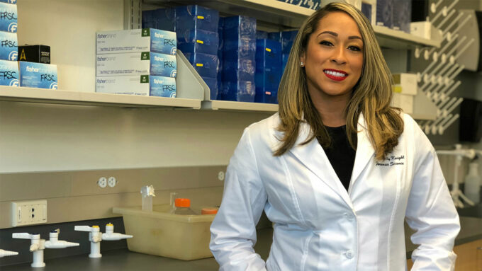 Kelly Knight is wearing a white lab coat. She is standing next to a green counter with various lab tools placed on top.