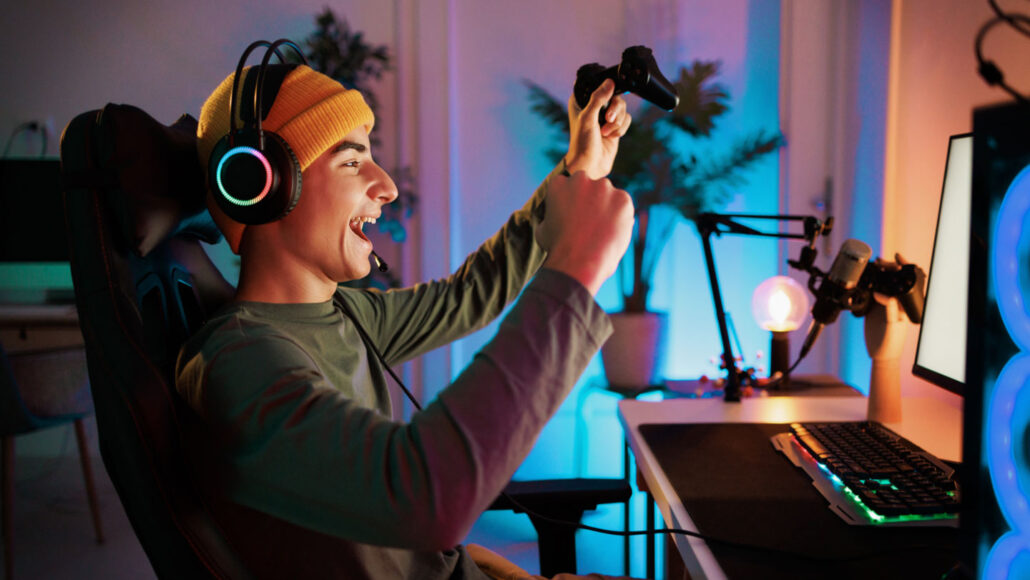 a boy sitting in front of a desktop computer and wearing headphones raises his hands and smiles in celebration