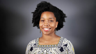 Civil Engineer Destenie Nock stands in front of a gray starburst background. She is a black woman with medium length, curly hair. She is wearing a tan blouse with black swirls and patterns.