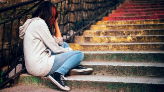 an upset teen turned away from the viewer sitting on an outdoor staircase