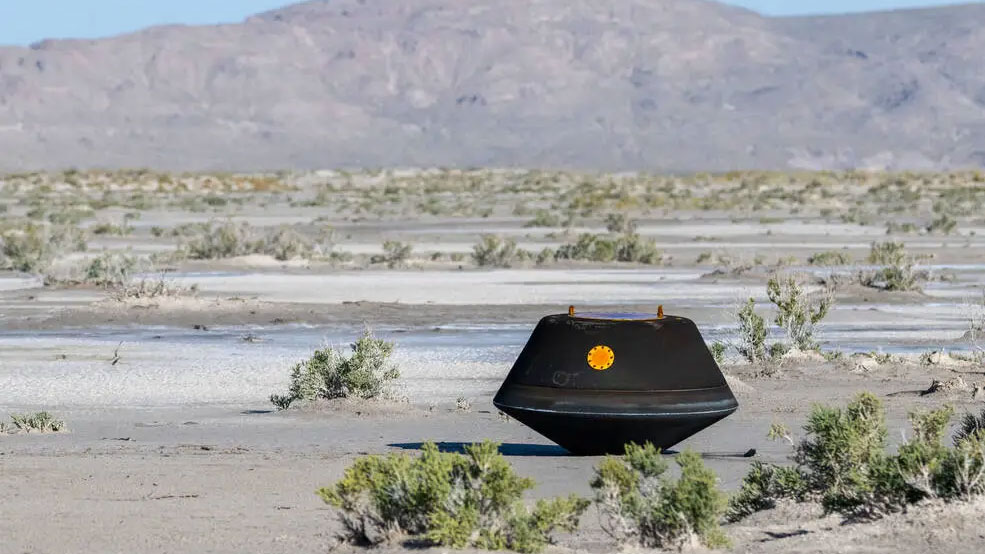 a black cylindrical capsule sits on the flat ground of a desert between tufts of shrubbery on a sunny day